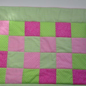Könnte beinhalten: Ein Patchwork-Quilt mit abwechselnden rosa und grünen Quadraten. Die rosa Quadrate haben ein Blumenmuster, die grünen Quadrate ein strukturiertes Design. Ein grün-weißes Vichy-Bordüre umrahmt den Quilt.