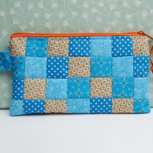 Könnte beinhalten: Ein gesteppter Beutel mit Patchwork-Design. Der Beutel hat Quadrate aus blauem, orangefarbenem und weiß gemustertem Stoff. Er hat einen orangefarbenen Reißverschluss und eine kleine Stoffschlaufe an der Seite. Der Beutel ist etwa 20 cm lang.