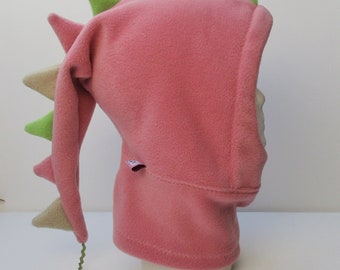 May include: Pink fleece dragon hooded hat with a green and beige spiky tail. The hat has a long tail that can be tied around the neck.