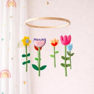 May include: A colorful felt flower mobile with a wooden ring. The mobile features various flowers in yellow, purple, orange, pink, and blue, with green stems and leaves. The mobile hangs in front of a sheer white curtain with rainbow and star designs.