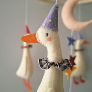 Puede incluir: Móvil infantil caprichoso con gansos de fieltro con sombreros de fiesta y pajaritas. Los gansos tienen picos naranjas y mejillas rosadas. Otras formas de fieltro incluyen una luna creciente y estrellas. El móvil está suspendido de un aro de madera.
