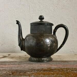 May include: A silver teapot with an ornate design and a fluted lid. The teapot has a rounded body and a curved spout. It is sitting on a wooden surface.