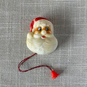 May include: A molded plastic Santa Claus head ornament featuring a red hat, a white beard, and blue eyes. A thin red string is attached to the bottom of the head, ending in a small red bell. The Santa face has a glossy finish and detailed texture.