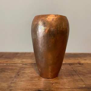 May include: A cylindrical copper vase with a hammered, textured surface. The metal displays a warm, metallic brown tone with subtle variations in color and a polished finish. The vessel has a slightly tapered base and a smooth, open top rim.