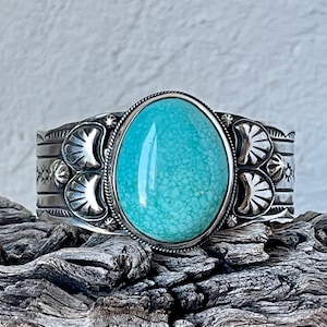 Może przedstawiać: Srebrna bransoletka z turkusowym kamieniem osadzonym w środku. Bransoletka jest ozdobiona misternymi srebrnymi zdobieniami i ma falisty wzór.