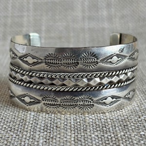 May include: A silver cuff bracelet featuring a multi-layered design with two wide bands displaying stamped sunburst and diamond patterns, separated by a central twisted rope wire band and a textured band with a repeating geometric diamond motif.