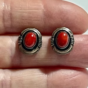 Puede incluir: Un par de pendientes de botón plateados, cada uno con una piedra ovalada roja vibrante. Las piedras están engastadas en un marco detallado y texturizado. Los pendientes se muestran sobre un fondo neutro.
