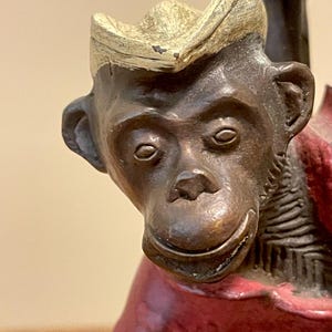 May include: A bronze-toned monkey figurine wearing a cream-colored hat and a dark red shirt. The monkey has a smooth, metallic finish with detailed facial features, including prominent ears, a small nose, and a subtle, closed-mouth smile.