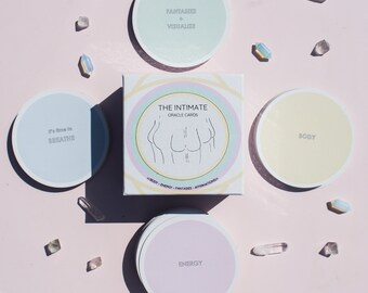 Könnte beinhalten: Ein Set von "The Intimate Oracle Cards" mit einer Box und vier kreisförmigen Karten. Die Box hat eine Strichzeichnung eines Körpers und die Worte "The Intimate Oracle Cards". Die kreisförmigen Karten haben Text wie "Body" und "Energy". Kristalle sind um die Karten verstreut.