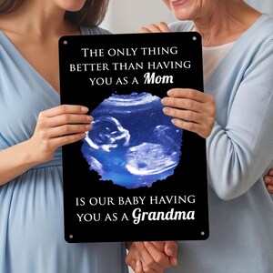 May include: A rectangular black metal sign featuring a central circular blue ultrasound image of a fetus. The text reads: THE ONLY THING BETTER THAN HAVING YOU AS A Mom IS OUR BABY HAVING YOU AS A Grandma. The sign has small holes in the corners.