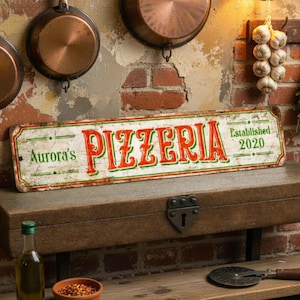 Può includere: Un'insegna rettangolare in metallo con finitura usurata che riporta il testo rosso e verde Aurora's PIZZERIA ed Established 2020. L'insegna è posizionata su una scatola di legno, accompagnata da una bottiglia d'olio, una ciotola di peperoncino e una rotella tagliapizza.