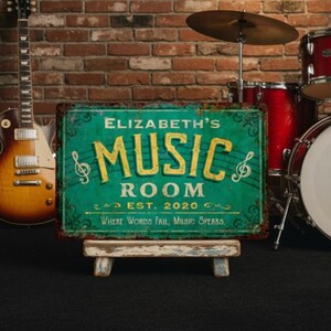 Puede incluir: Un letrero metálico turquesa y amarillo que dice "Elizabeth's Music Room" con notas musicales, colocado sobre un pequeño caballete de madera. Una guitarra y una batería están en el fondo contra una pared de ladrillos.