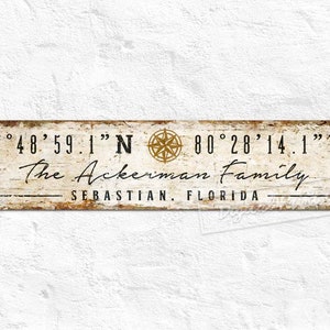 May include: A white wooden sign with black text and a compass design. The sign reads "27°48′59.1″N 80°28′14.1" W The Ackerman Family SEBASTIAN, FLORIDA"