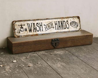 May include: A rectangular metal sign with a distressed white finish and black text reading WASH YOUR HANDS, featuring a graphic of a faucet and a bar of soap, resting atop a long, dark brown wooden box with a central metal latch and keyhole.