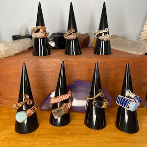 May include: A collection of handcrafted wire-wrapped rings displayed on black cone-shaped stands. The rings feature various gemstones and beads in colors like blue, green, orange, and pink. The rings are showcased on a wooden surface.
