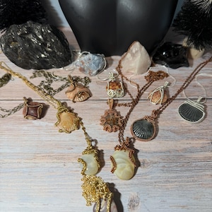 May include: A collection of handcrafted necklaces featuring various gemstones and shells, each uniquely wire-wrapped. The necklaces showcase a range of colors, including brown, gold, and white, with different chain styles. The jewelry is displayed on a light wooden surface.