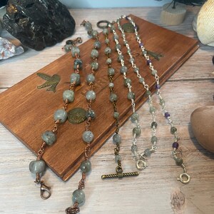 May include: Four beaded bracelets with various designs. The bracelets feature gray and silver beads, copper and silver wire, and decorative clasps. They are displayed on a wooden box with a bird design.