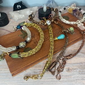 Peut inclure: Une sélection de bracelets faits à la main dans divers styles et couleurs, dont argent, or et cuivre. Certains bracelets présentent des perles, des breloques et des motifs en fil de fer. Les bracelets sont exposés sur une boîte en bois.