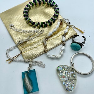 May include: A set of jewelry featuring a turquoise ring, a shell necklace, a silver chain necklace with a blue resin pendant, a green and purple beaded bracelet, and a silver key chain with a clear resin charm with silver glitter.
