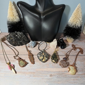 Peut inclure: Plusieurs colliers artisanaux avec des pierres précieuses, présentés sur une surface en bois. Les pendentifs sont ornés de pierres vertes, violettes et claires, avec un fil de cuivre. Un buste de mannequin noir et des arbres décoratifs sont en arrière-plan.