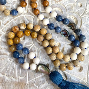 K&ouml;nnte beinhalten: Eine mehrstr&auml;ngige Mala-Halskette mit Holz-, wei&szlig;en und blauen Perlen. Die Halskette hat einen Quasten aus blauem Stoff und einen Muschel-Anh&auml;nger.