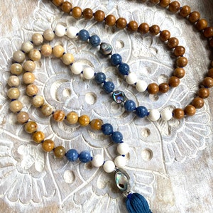 K&ouml;nnte beinhalten: Eine mehrstr&auml;ngige Mala-Halskette mit einem blauen Quastenanh&auml;nger. Die Halskette besteht aus Holz, wei&szlig;en und blauen Steinen und hat Akzente aus Abalone-Muschel.