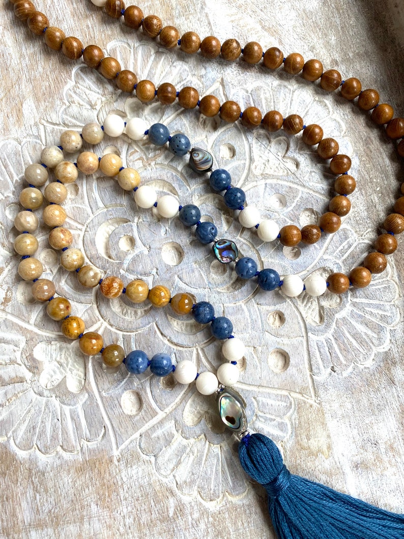 K&ouml;nnte beinhalten: Eine mehrstr&auml;ngige Mala-Halskette mit einem blauen Quastenanh&auml;nger. Die Halskette besteht aus Holz, wei&szlig;en und blauen Steinen und hat Akzente aus Abalone-Muschel.