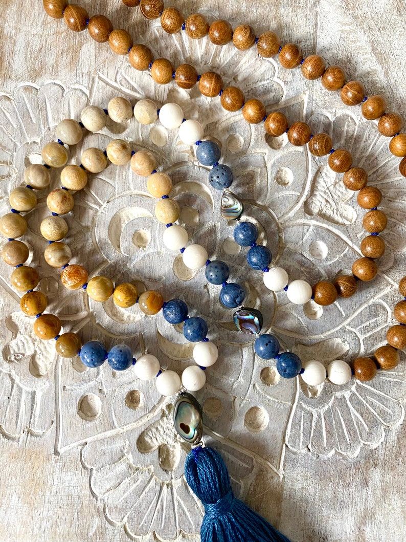 K&ouml;nnte beinhalten: Eine blaue, wei&szlig;e und braune Perlenkette mit einem blauen Quasten. Die Kette besteht aus Holz-, wei&szlig;en Stein- und blauen Steinperlen. Die Quaste ist aus blauem Stoff.