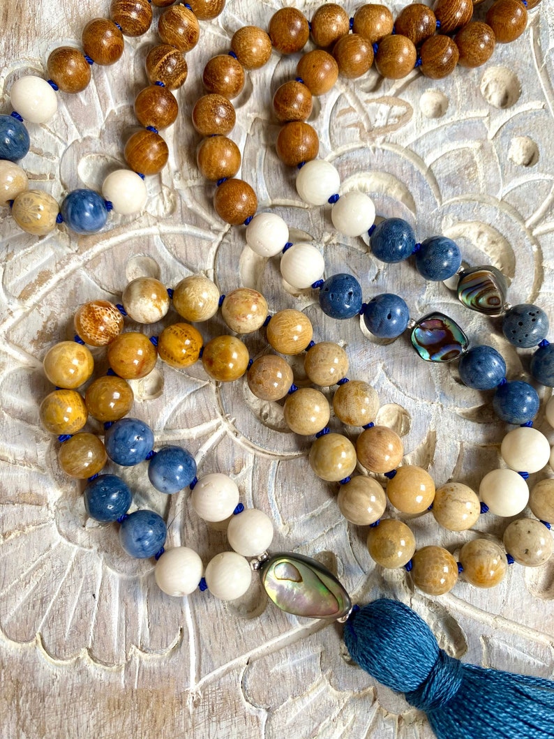 K&ouml;nnte beinhalten: Eine mehrfarbige Perlenkette mit einem blauen Quasten. Die Kette besteht aus Holz-, Stein- und Muschelperlen. Die Perlen sind in einem Farbverlauf angeordnet, beginnend mit Braun, dann Gelb, dann Blau und schlie&szlig;lich Wei&szlig;. Die Quaste ist aus blauem Stoff.