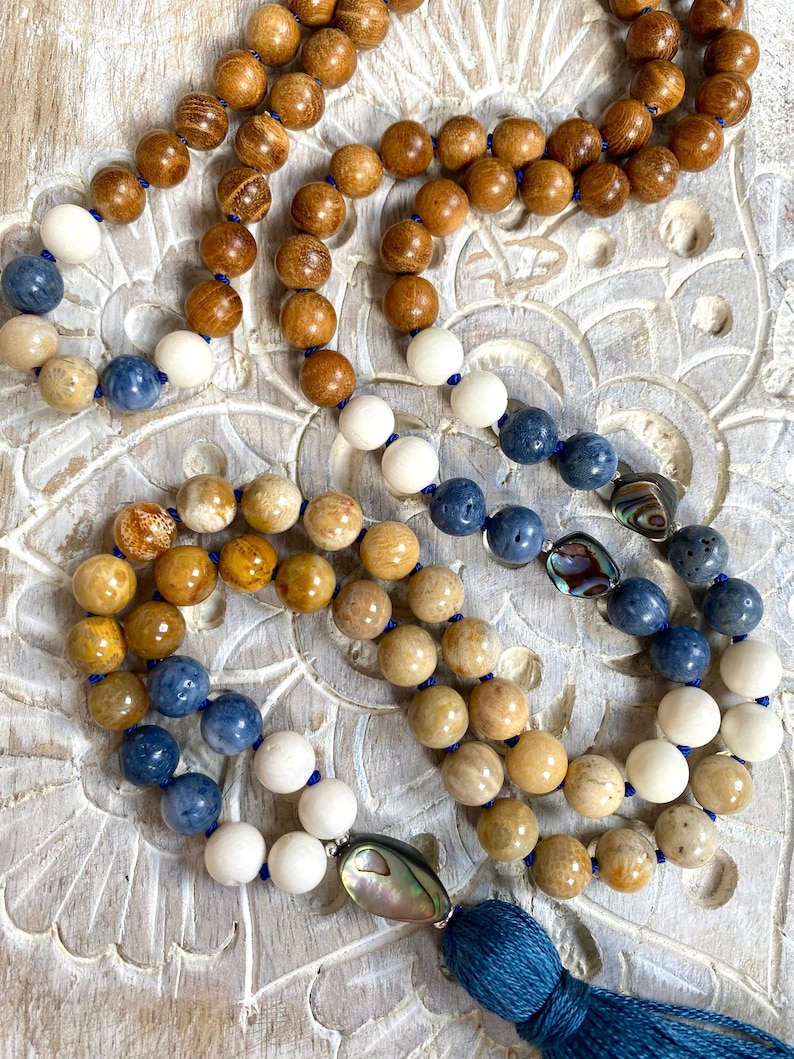 K&ouml;nnte beinhalten: Eine mehrstr&auml;ngige Mala-Halskette mit Holz-, wei&szlig;en und blauen Perlen. Die Halskette hat einen Quasten aus blauem Stoff und einen Muschel-Anh&auml;nger.