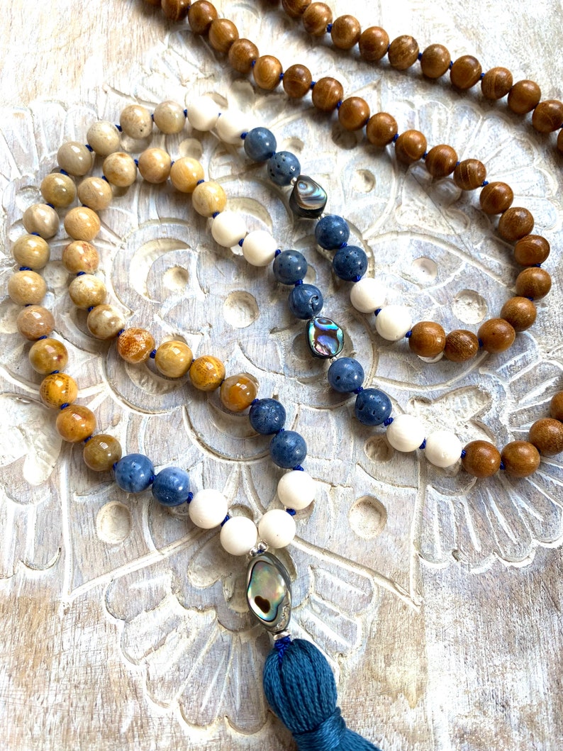 K&ouml;nnte beinhalten: Eine mehrfarbige Perlenkette mit einem blauen Quastenanh&auml;nger. Die Kette besteht aus Holz-, wei&szlig;en, blauen und braunen Perlen. Die Perlen sind in einem Farbverlauf angeordnet. Die Kette hat einen Muschel-Anh&auml;nger am unteren Ende.