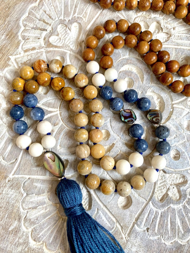 K&ouml;nnte beinhalten: Eine mehrstr&auml;ngige Mala-Halskette mit einem blauen Quastenanh&auml;nger. Die Halskette besteht aus Holz-, versteinertem Korallen-, wei&szlig;en Stein-, blauen Stein- und Lavakugeln. Die Perlen sind mit Abalone-Muschel-Akzenten durchsetzt.