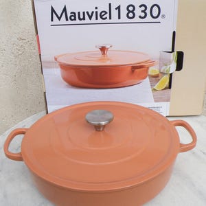 May include: A Mauviel 1830 cast iron Dutch oven with a lid. The oven is orange and has two handles. The lid has a silver knob.