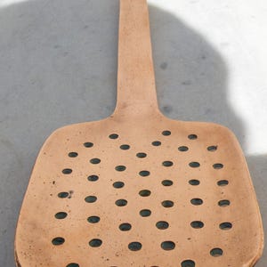 Op de afbeelding: Een koperen geperforeerde lepel met een lange steel en een platte, ovale kop met gelijkmatig verdeelde ronde gaten.