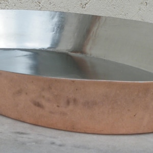 May include: Oval copper roasting pan with a shiny silver interior. The pan has a copper exterior and a brass handle. The pan is designed for cooking and baking.