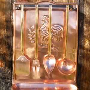Peut inclure: Ensemble d'ustensiles de cuisine en cuivre avec un relief de coq. L'ensemble comprend une spatule, une fourchette, une cuillère et une louche, tous suspendus à un support mural en cuivre. Les ustensiles ont des manches en laiton.