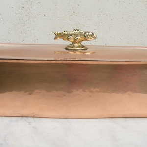 May include: A large, oval copper roasting pan with a lid. The lid has a decorative brass fish finial. The pan has two brass handles shaped like fish.