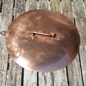 Könnte beinhalten: Ein runder Kupfertopfdeckel mit Griff und Ring an der Seite. Der Deckel hat eine glänzende Oberfläche und liegt auf einem verwitterten Holzhintergrund. Der Griff ist am Deckel vernietet.