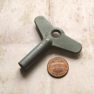 May include: A gray metal clock key with a T-shaped handle and a circular hole. A copper one-cent coin is next to the key for size comparison. The key is likely for winding a mechanical clock or other device.