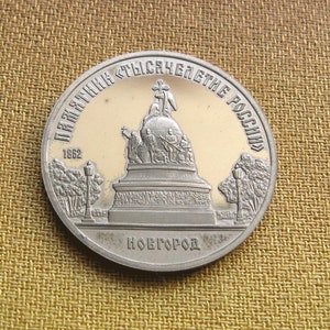 Puede incluir: Una moneda de plata que conmemora el Milenio de Rusia con una representación del Monumento al Milenio de Rusia en Novgorod. La moneda tiene la inscripción "Памятник Тысячелетие России" (Monumento al Milenio de Rusia) y "Новгород" (Novgorod).