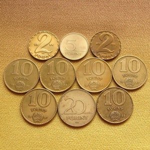 May include: A collection of vintage Hungarian Forint coins, featuring denominations of 2, 5, 10, and 20. The coins are gold-colored and arranged on a textured, golden-brown surface. The coins display the word "FORINT" and the year of minting.