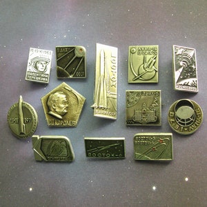 Könnte beinhalten: Eine Sammlung von 12 silberfarbenen Metallstiften mit Weltraumforschungsmotiven. Die Stifte zeigen Raketen, Astronauten und den Mond. Die Stifte sind in einem Rastermuster auf einem dunkelgrauen Hintergrund angeordnet.
