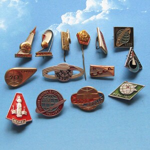 Può includere: Una collezione di 13 spille smaltate vintage con temi di esplorazione spaziale. Le spille sono di vari colori, tra cui rosso, blu, verde, oro e argento. Alcune delle spille mostrano il testo "URSS" e "CCCP".