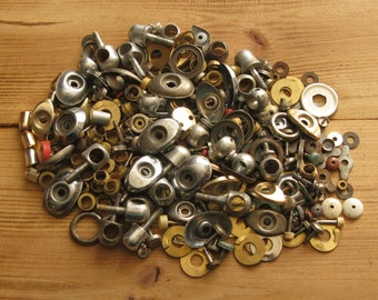 May include: A large pile of assorted metal hardware, including screws, nuts, washers, and other small metal parts. The hardware is mostly silver and gold in colour, and is arranged in a random pattern on a wooden surface.
