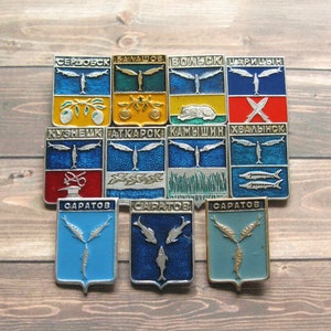 May include: A collection of vintage enamel pins featuring various city crests. Each pin has a rectangular or shield shape with a silver emblem and text. The pins display a variety of colors, including blue, yellow, red, and green, and are arranged on a wooden surface.