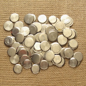 Op de afbeelding: Een grote stapel zilveren horlogebodems, sommige met een verhoogde rand, op een bruine jute achtergrond.