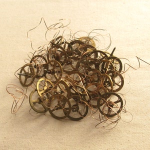 May include: A collection of small, brass clock gears and copper springs scattered on a beige surface. The gears vary in size and shape, with intricate details. The copper springs are coiled and add a contrasting color and texture to the arrangement.