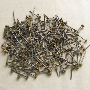 Op de afbeelding: Een grote stapel kleine, zilverkleurige metalen staven met gouden tandwielen aan het uiteinde. De staven liggen verspreid op een wit oppervlak.