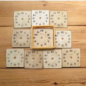 Pode incluir: Nove mostradores de relógio vintage com números pretos e fundo branco. Os mostradores de relógio estão dispostos em três filas de três. Os mostradores de relógio são todos ligeiramente diferentes, alguns têm um pequeno furo no centro e outros um furo maior. Os mostradores de relógio são todos de papel e têm um aspeto ligeiramente envelhecido.