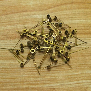 May include: A pile of gold-coloured clock hands, some with small metal rings at the end, on a light brown wooden surface.